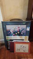 Overall view of multiple framed and unframed artworks stacked against a wall, showing variety and size relation, including visible floral print and red-framed drawing