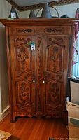Front view of a large Henredon carved wood armoire with floral and fruit carvings on the doors and drawers. Keys visible on doors. Positioned against wall with household items around.