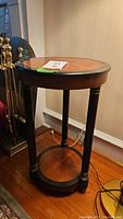 Front view of round side table showing inlaid wood look top, three black legs with gold trim and round bottom shelf.