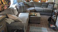 Three-piece gray sectional arranged in L-shape against brick floor with ottoman and throw pillows