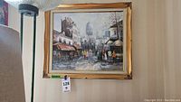 Frontal view of the framed oil painting hanging on a wall under a lamp, showing street scene and signature.