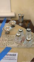 Wide shot of all items on table showing 4 crystal salt shakers, 2 silver salt servers with spoons, and 2 silver toasting cups