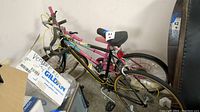 Two Huffy bikes side by side, one black with yellow striping, the other pink Miss Huffy, located indoors near boxes and garage items.
