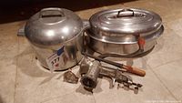 Photo showing large covered aluminum pot, oval roaster with lid, hand-crank meat grinder with wooden handle, and an oven thermometer on the floor.