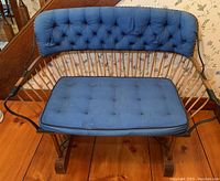 View of antique wooden bench from the front showing tufted blue cushioned backrest and removable seat cushion on wooden frame with vertical spindles and curved rocker base.