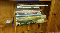 Top view of a stack of art books on a wooden shelf, titles visible include ones about Da Vinci, Brooklyn Museum, Chinese Art, and American Art.