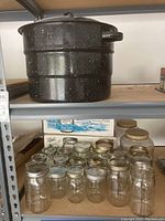 Enamel speckled black canning pot sitting on metal shelving above multiple glass jars with metal lids and a box.