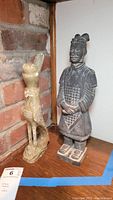Photo of ceramic Chinese warrior statue and carved stone bird sculpture side by side on a shelf.