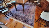 Full view of the Reubens wool area rug situated under a wooden dining table and chairs on hardwood flooring. The rug displays a traditional Flemish pattern with a border and central medallion in navy, beige, and muted red colors.