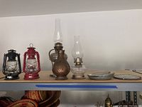 Photo showing two metal framed lanterns (black and red), a copper-colored metal oil lantern with glass hurricane, a glass oil lamp, and two metal trays on a shelf.
