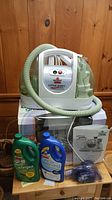 Front view of the Bissell Little Green Machine with hose coiled, two cleaning solution bottles, user guide, and an accessory tool on a wooden table.