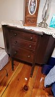 Front view of vintage wooden sewing chest covered with a lace doily, showing a clock and lamp on top.