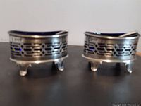 Pair of sterling silver salt cellars with pierced geometric design and three legs, placed side by side on black surface.
