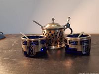 Two cobalt blue glass salt cellars with silver holders and spoons, silver plated mustard pot with lid and spoon on a dark surface.