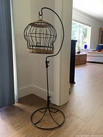 Full view of bird cage on stand placed near a wall and window, showing wire cage with woven rattan top and bottom and curved metal stand