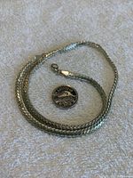 Silver heavy chain necklace arranged on a light towel surface with a US quarter for scale, showing reflective and woven link design.