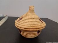 Full view of small conical lidded basket showing woven natural and dyed fibers with geometric patterns