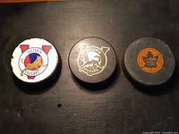 Three vintage hockey pucks: Victoria Cougars puck with white logo, Victoria Cougars puck with gold logo, and Victoria Maple Leafs puck with orange maple leaf logo, shown on black surface.