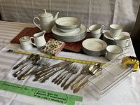 Full view of dishes, cups, cutlery, teapot, creamer, sugar bowl, placemats, napkins, and cutlery organizers arranged on a table with tape measure and note describing contents.