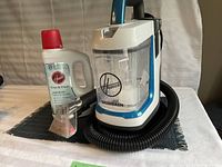 Clear image of Hoover Power Dash spot cleaner unit with hose and cleaning attachment alongside bottle of Hoover Free & Clean cleaning formula on a black mat background.
