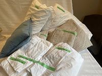 Photo showing white and beige bed linens, pillows, and blue throw cushion with labels on some items for identification.