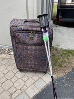 Full view of paisley patterned rolling suitcase with two walking poles leaning against it on paved ground with grass and building wall in background.
