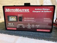 Front view of MotoMaster battery charger showing brand name, model, analog ammeter, 10/2A charging selector switch, and usage instructions.