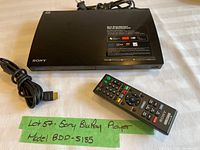 Sony BDP-S185 Blu-Ray Disc/DVD Player with remote control and HDMI cable on a white surface. Front view showing power cable and USB port.
