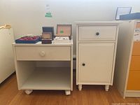 Two white laminated nightstands side-by-side, showing the shorter one with drawer and open shelf on wheels and the taller one with drawer and cabinet door.