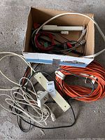 Photo of assorted extension cords and power bar, showing cords coiled and placed inside and outside of a cardboard box on concrete floor