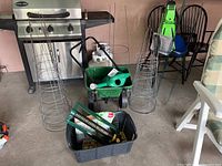 Photo of three metal tomato cages, wheeled green spreader, black plastic bin with gardening tools, and green watering can on concrete floor in garage area.