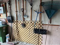 Multiple garden tools hung on wall including shovels, rakes, pitchfork, axe, broom, showing full lot contents