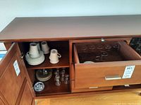 Wide view of cabinet with white Mikasa mugs and saucers stacked on shelves and German thin glasses in drawer.