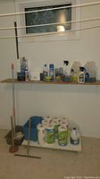 Photo of cleaning supplies including mop, mop buckets, paper towels, and various cleaning chemicals on shelf and floor in basement room with window