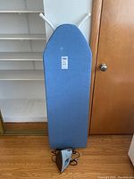 Blue fabric covered folding ironing board upright on floor with iron placed in front at base.