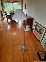 Floor lamp with white fabric shade and metal base shown in a room with wooden floor, dining table and framed art leaning against wall in background.