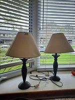 Picture of two metal base table lamps with beige fabric shades placed on a white surface in front of window blinds.