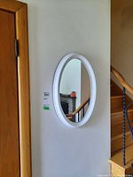 Framed oval mirror on a white wall, near wooden staircase and black metal railing, reflecting part of a fireplace mantel with orange candle and decor.