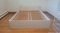 White Ikea queen size bed frame assembled against a wall on light hardwood floor, showing full length and width with visible pine slats.
