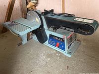 Side angled view showing Delta belt and disc sander with work table and power cord on concrete floor near wall.