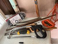 Various saws including a large hand saw with an orange handle, two hacksaws, and older hand saws with wooden handles alongside a black and orange hose with adaptor and copper pipe.