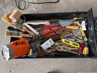 Overhead photo of the open box containing paint brushes, cutters, wrenches, pliers, utility knife, measuring tape, putty knife, and orange flashlight