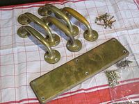 Photo of five solid brass door handles with curved design and round base, five rectangular solid brass push plates, and two bags with original brass screws placed on a red and white cloth.