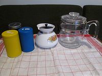 View of coffee pot, teapot, and three tea containers on a cloth surface, showing lot content