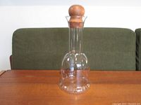 Full view of the clear glass decanter with its rounded teak stopper on top, placed on a wooden surface with a green cushioned background.