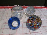 All four glass paperweights on a cloth background: clear hollow apple, clear bubble-filled sphere, blue Caithness with clock, colorful millefiori pattern paperweight.
