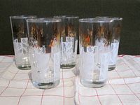Set of five highball bar glasses standing on cloth with white and red grid pattern, showing clear glass with gold and white motifs.