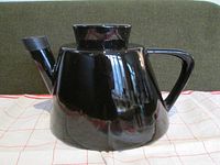 Black ceramic teapot placed on a cloth with checkered pattern, showing full side profile including handle and spout.