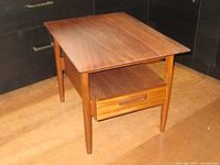 Front three-quarter view showing solid teak top, single drawer-shelf and tapered legs