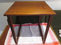 Front view showing the square teak tabletop and finger jointed legs attached by screws, placed on a red and white checkered cloth.